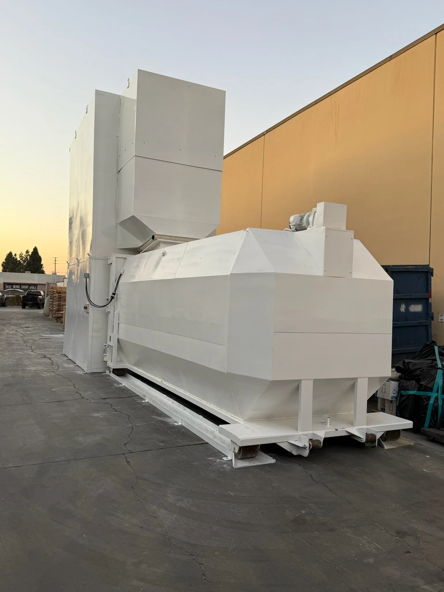 Large white industrial equipment or container sitting outdoors on a paved surface, with a beige building and a few trees in the background during sunset or early evening.
