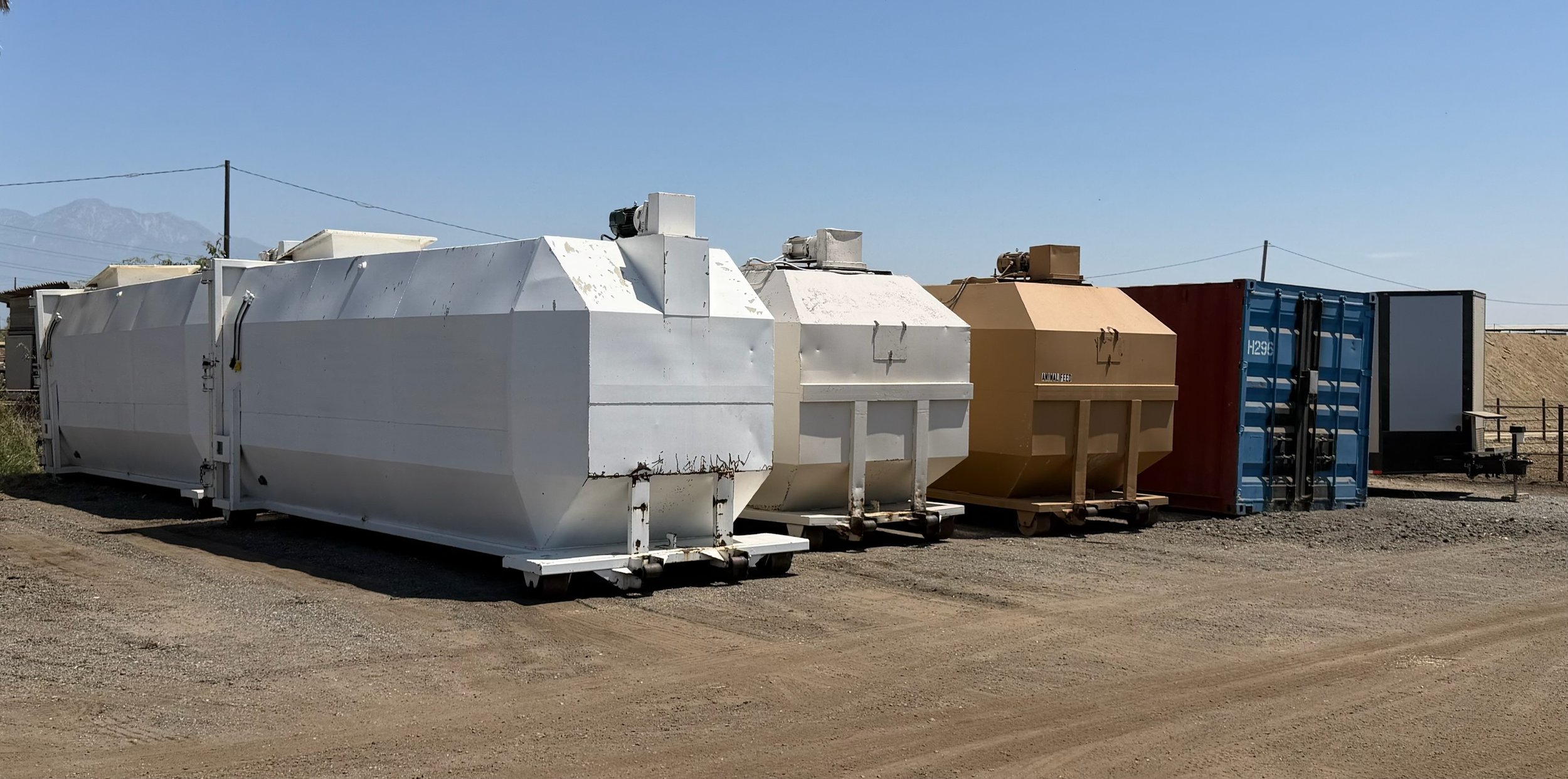 Various large collection of waste containers, including white, beige, blue, and red, placed outdoors on a dirt ground with a clear blue sky.