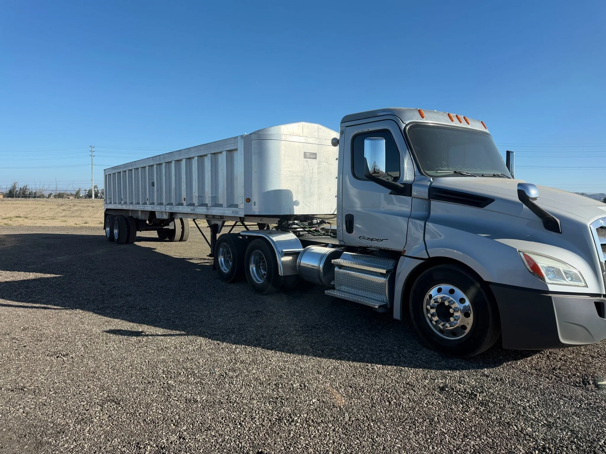 White semi-truck with a long flatbed trailer parked on gravel, under a clear blue sky with power lines in the background.