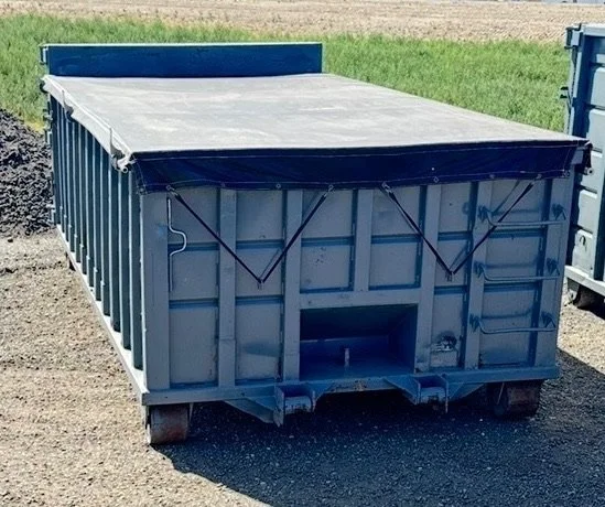 A large industrial gray shipping container with a partially open black tarp covering the top, placed on a gravel surface outdoors.