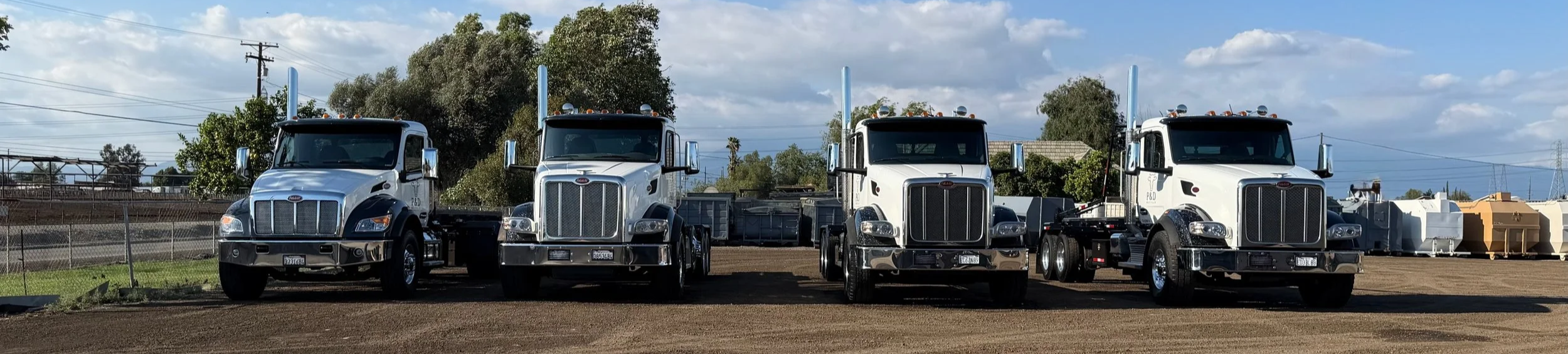 Four semi-trucks with black and white cabs parked side by side on dirt ground, with a background of trees, power lines, and industrial containers under a partly cloudy sky.