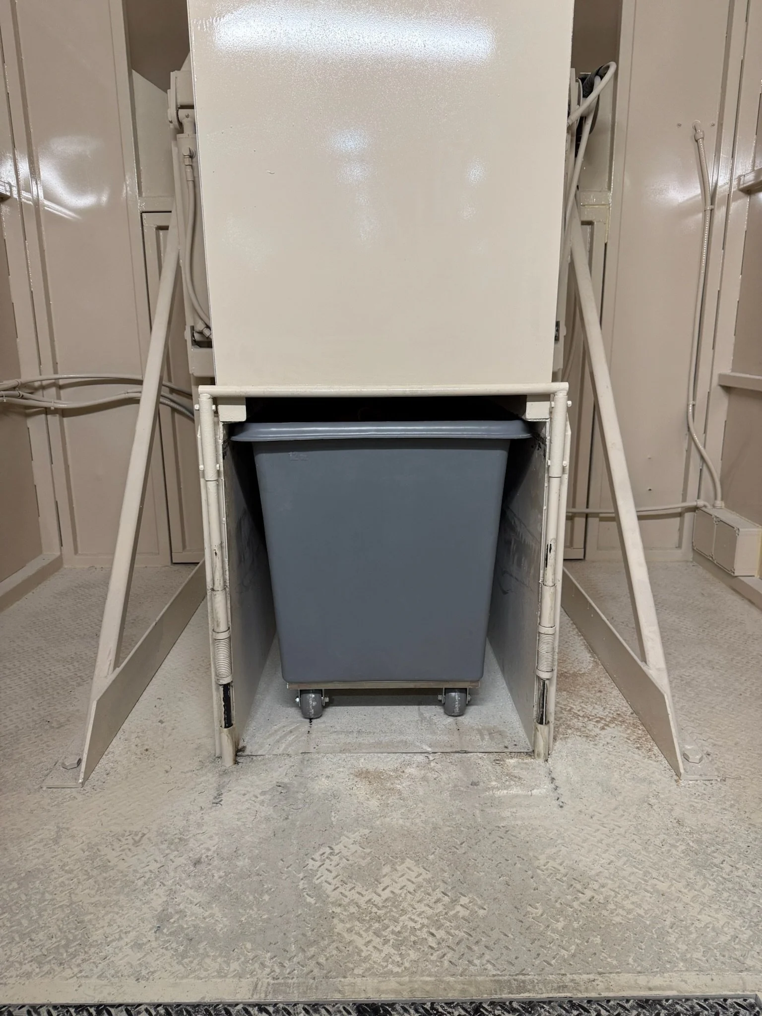 A laundry room with a white dryer and a gray laundry bin underneath it, on a white metal stand, with beige walls and a textured beige floor.