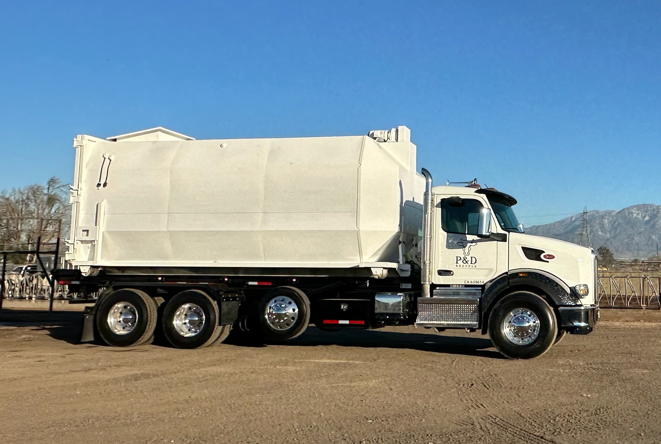 A white delivery truck with a refrigerated cargo box parked on a dirt lot, with mountains and power lines in the background, under a blue sky.