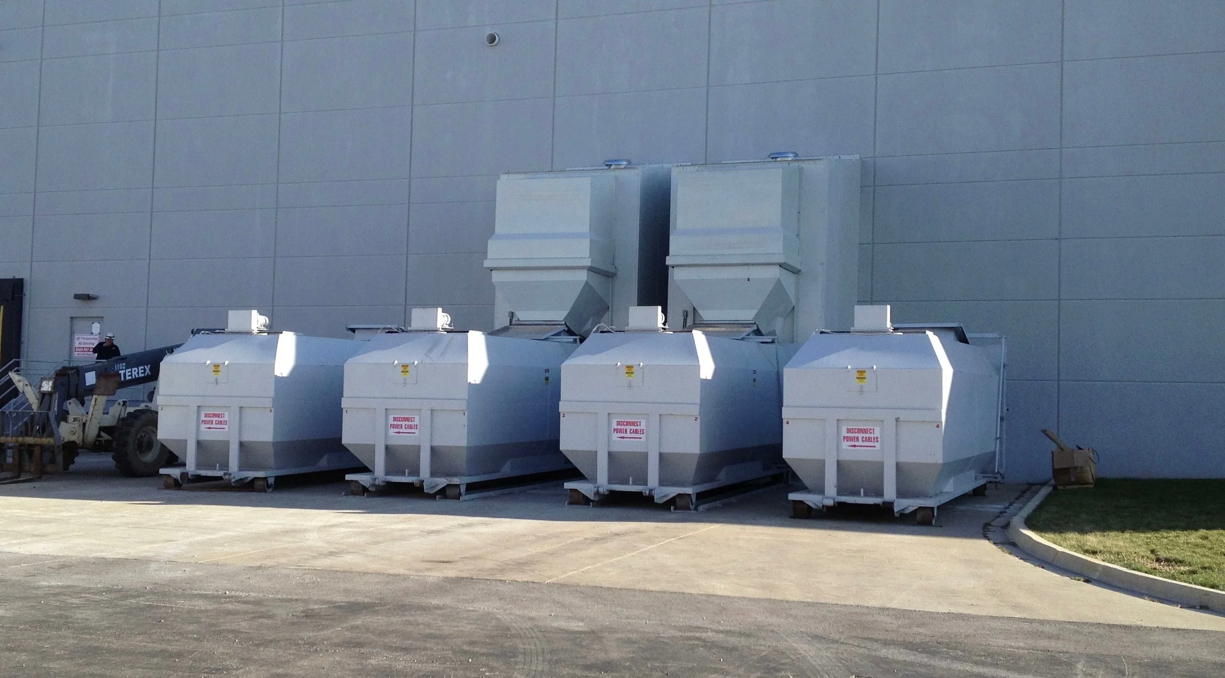 Large industrial air handling units or ventilation systems are positioned outside a building, with one worker standing nearby.