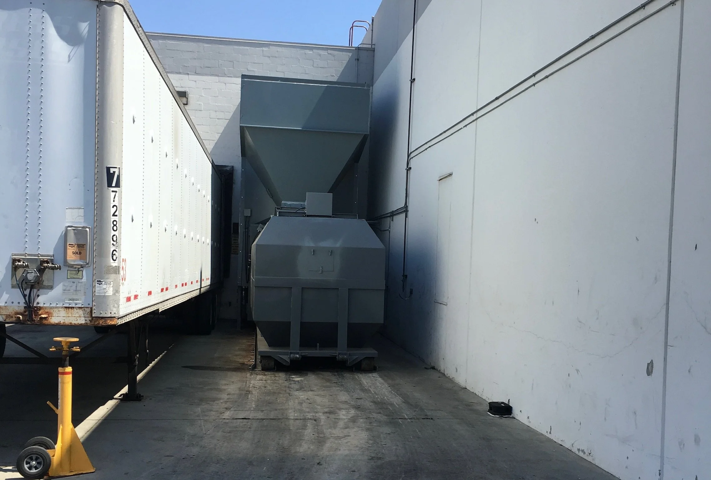 An industrial alleyway with a white building on the right and a truck on the left. HVAC units and ducts are visible; a yellow safety cone is in the foreground.