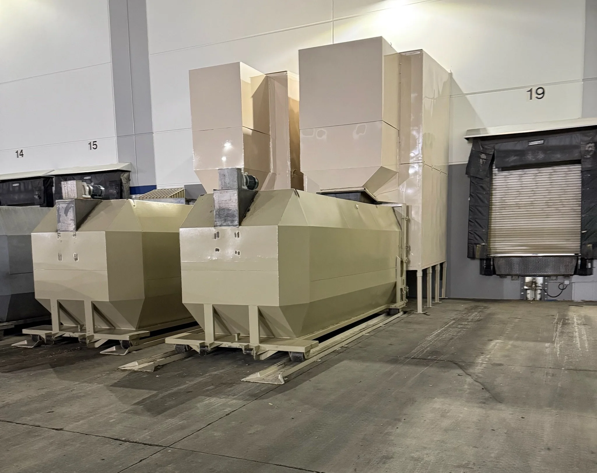 Industrial setting with large beige machinery or ventilation systems on a concrete floor, wall with numbered sections, and a closed roll-up door.