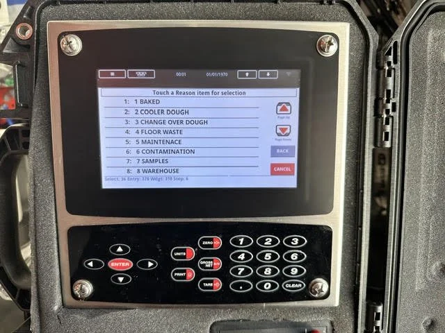 A touchscreen control panel with a list of options for selecting reasons, including baked, cooler dough, change oven dough, floor waste, maintenance, contamination, samples, and warehouse; with navigation buttons below.