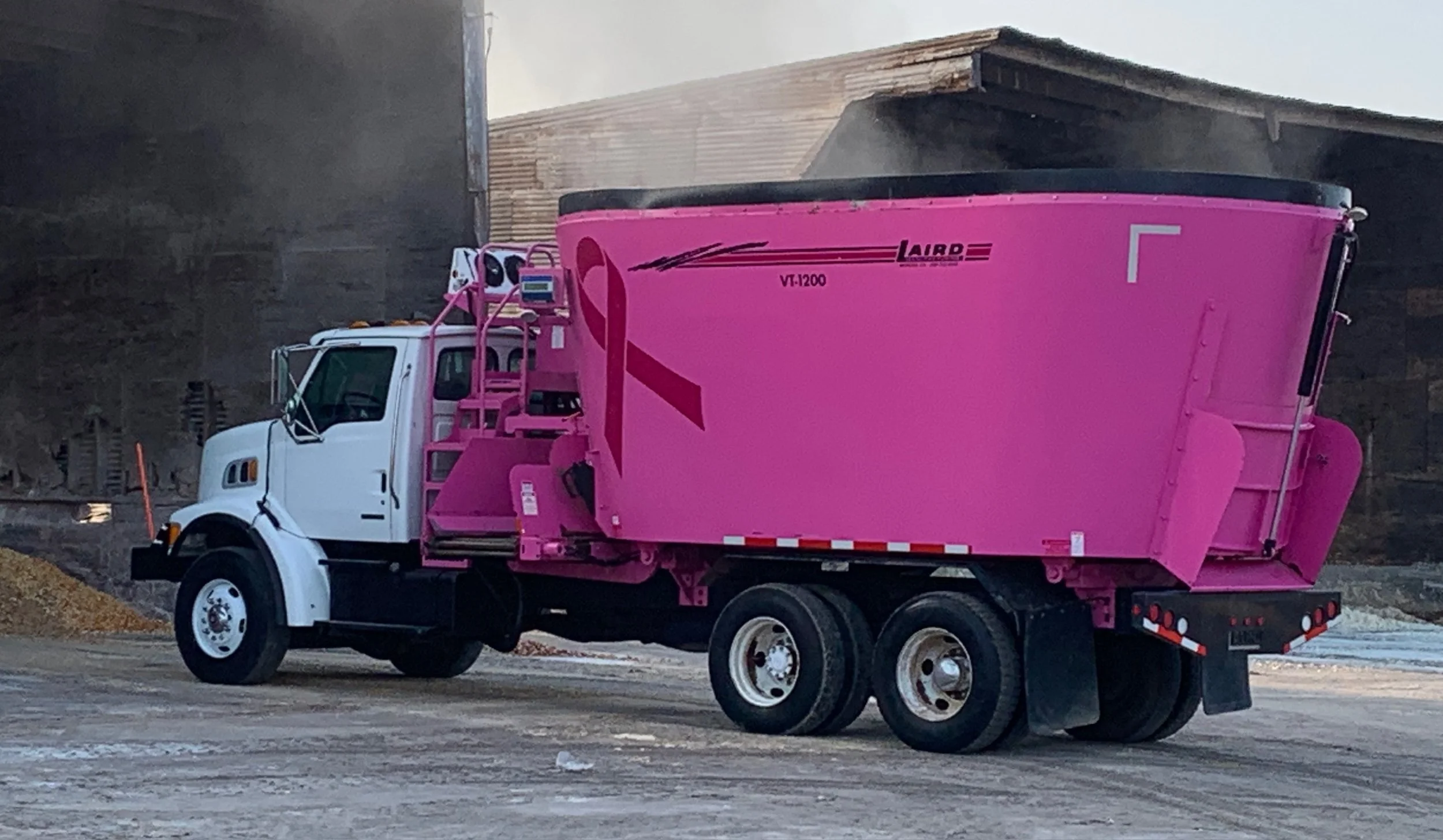 A white dump truck with a pink container on a construction site. The background shows a dirt mound and the sky.