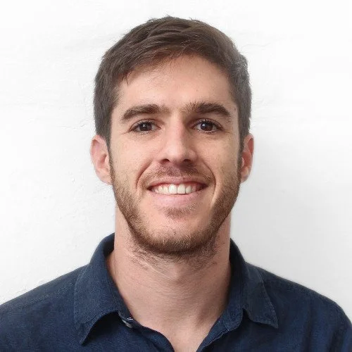 Portrait of a smiling man with short brown hair and a light beard, wearing a blue shirt against a white background.