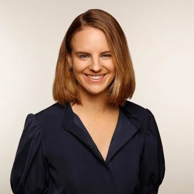 Smiling woman with shoulder-length hair wearing a dark blouse against a plain background.