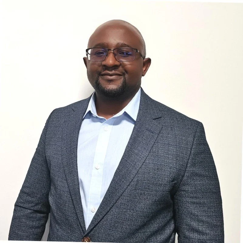 Man wearing a gray suit jacket and blue shirt, standing against a white background.