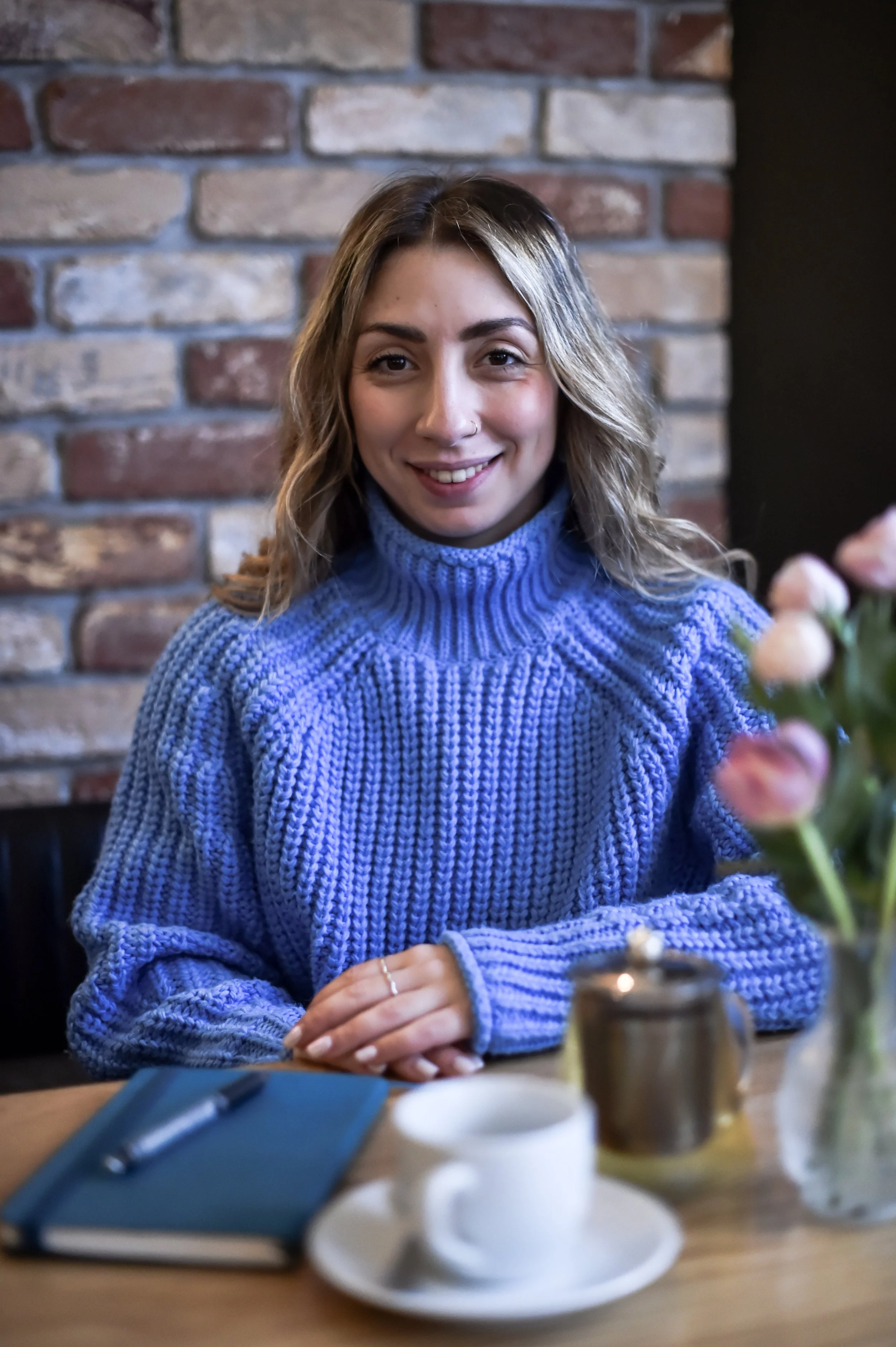 A woman with wavy, shoulder-length hair smiling at the camera while sitting at a table in a cozy café with a brick wall background. She is wearing a blue sweater and has a nose ring.