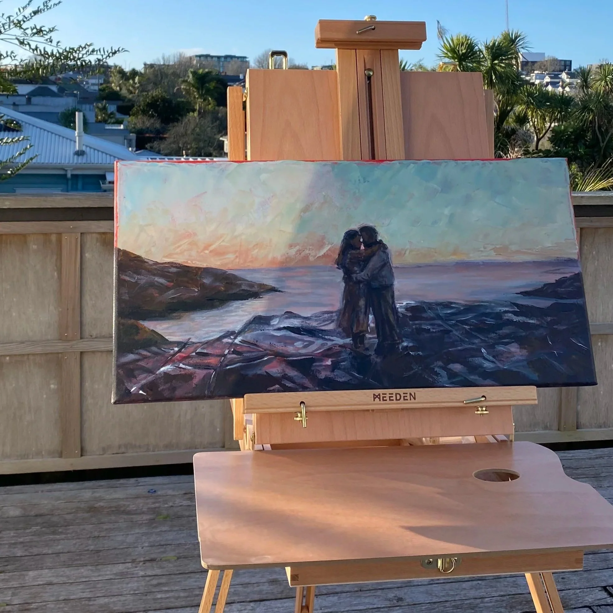 A painting on an easel depicting a couple kissing on a rocky beach during sunset, with trees and buildings in the background in New Zealand.