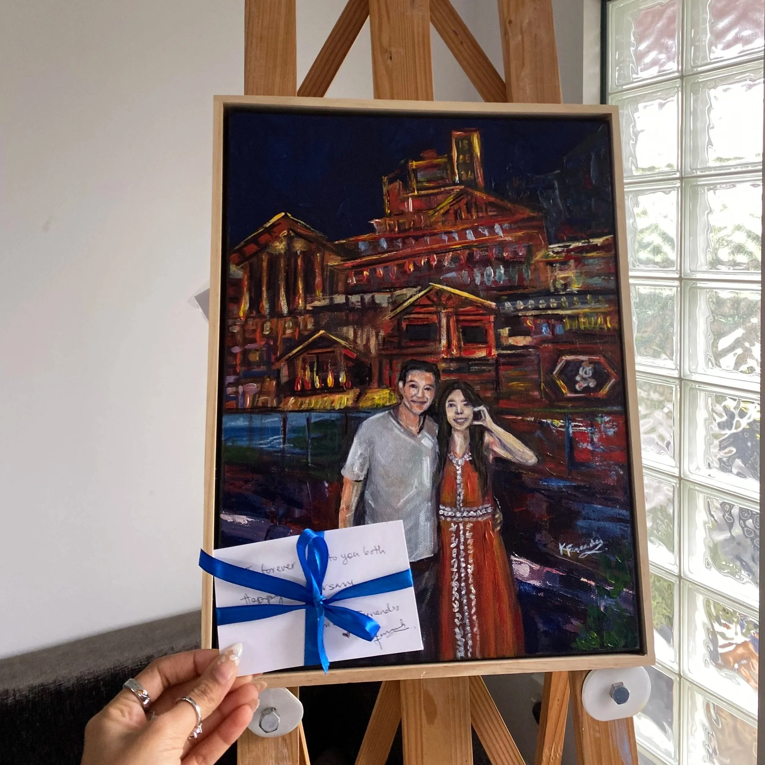 A painting of a couple standing in front of a large, brightly lit building, possibly a historic site or landmark, with a note attached to the frame tied with a blue ribbon.