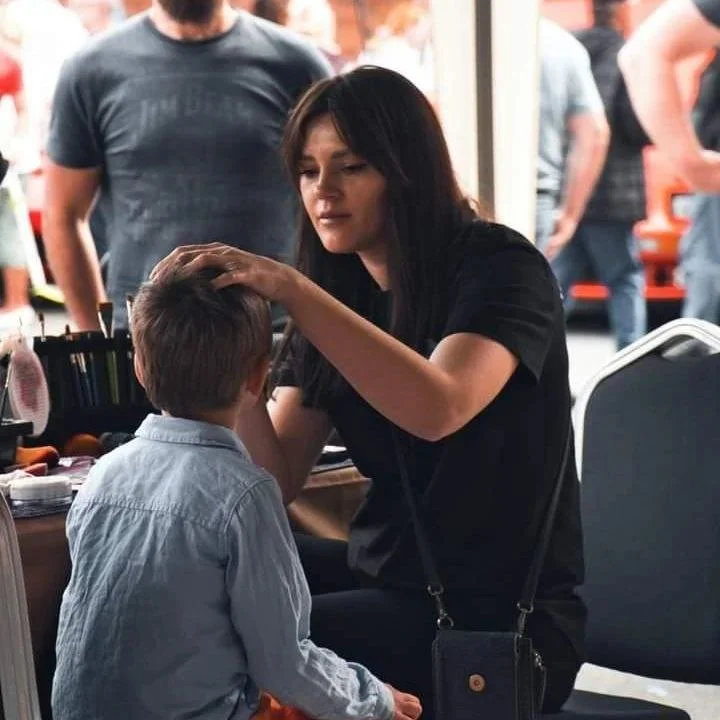 A woman is applying makeup or face paint to a young boy's face at an event, with other people in the background.