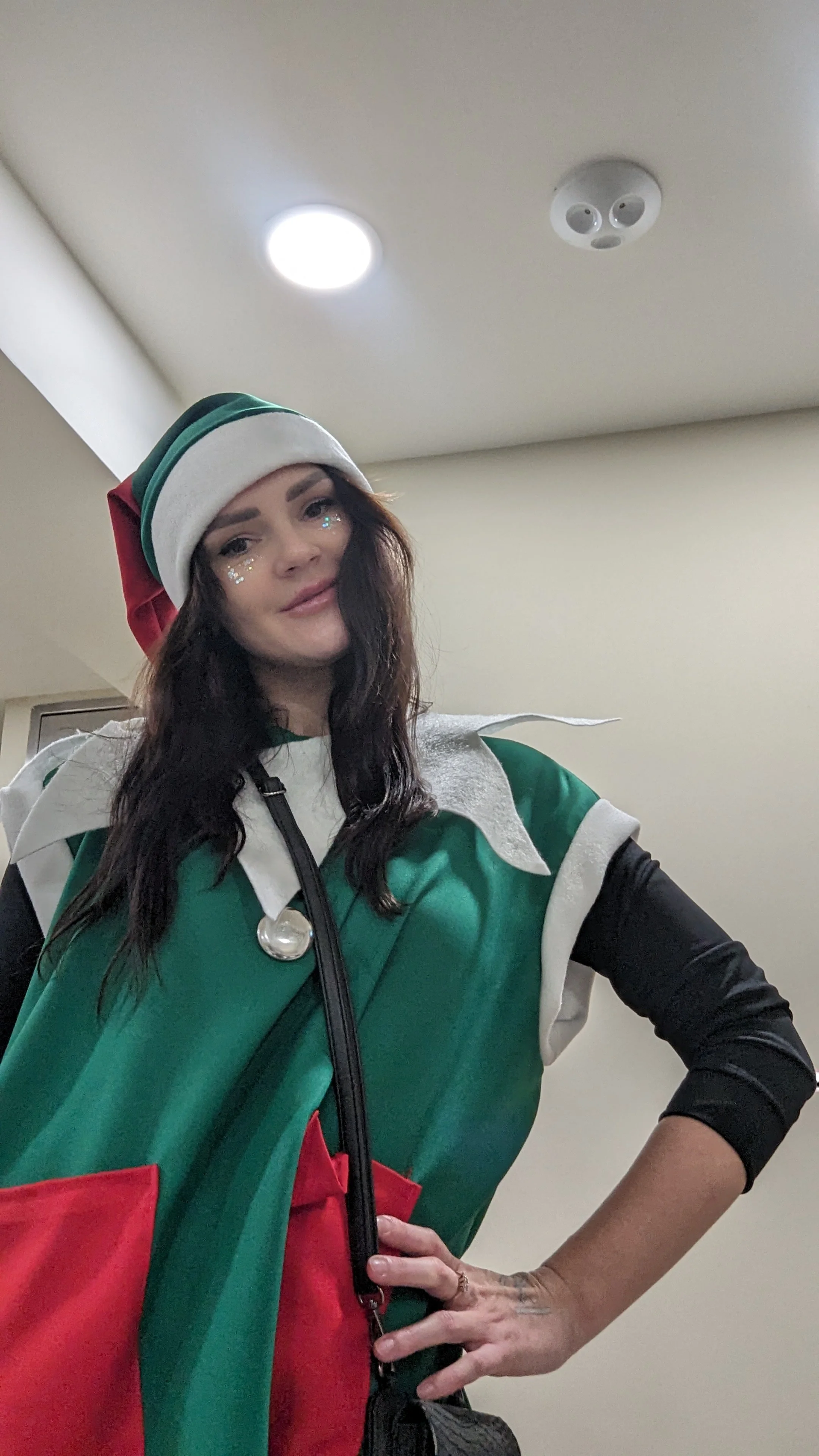 A woman dressed in an elf costume with a green shirt, white collar and shoulder accents, red pocket, black long-sleeve shirt underneath, and a green, white, and red Santa hat. She has long dark hair, glitter makeup around her eyes, and a hand on her hip, standing indoors against a plain wall.