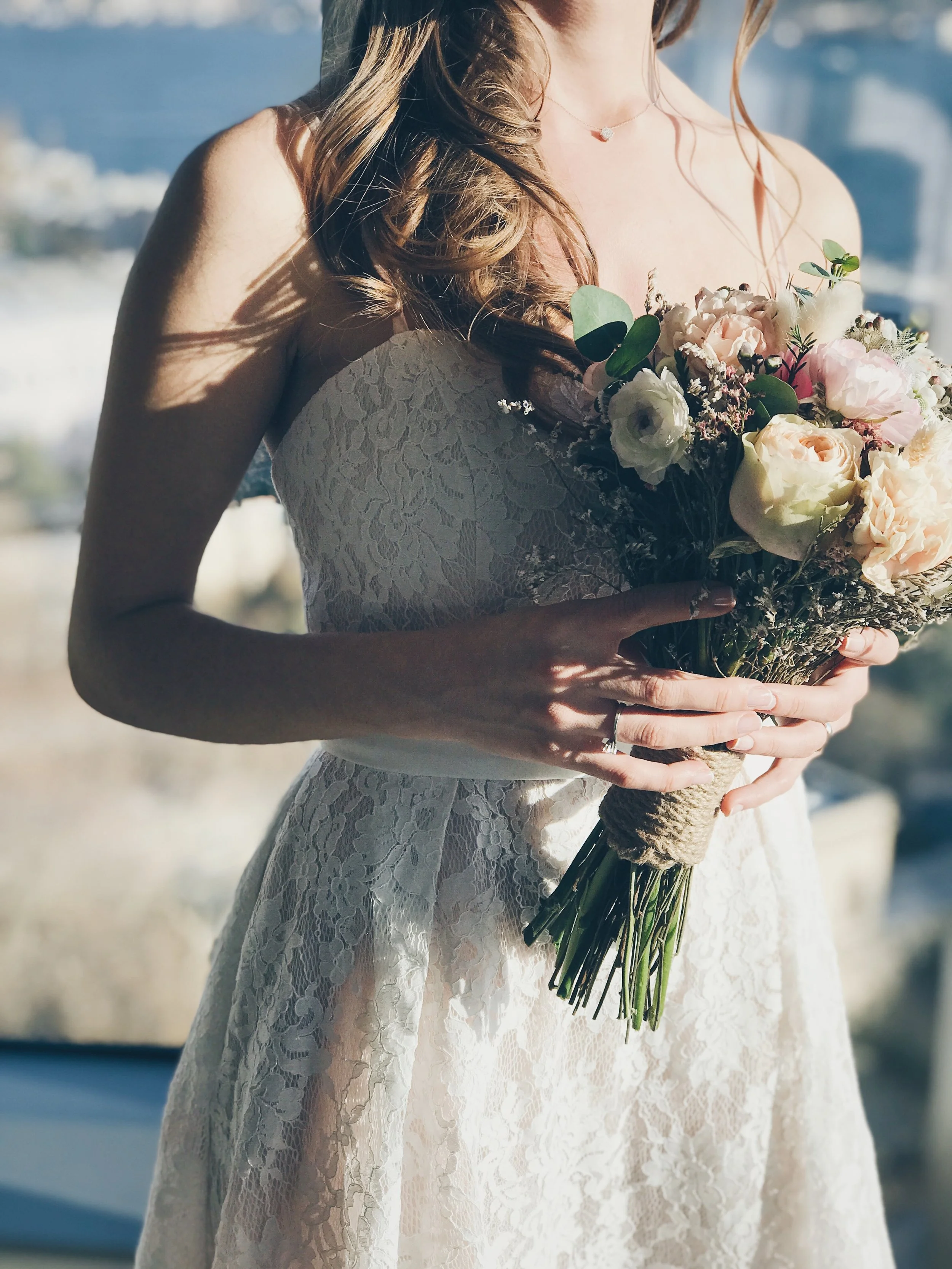 Close-up van een bruid in een kanten trouwjurk met een boeket bloemen, met een kustlijn op de achtergrond.
