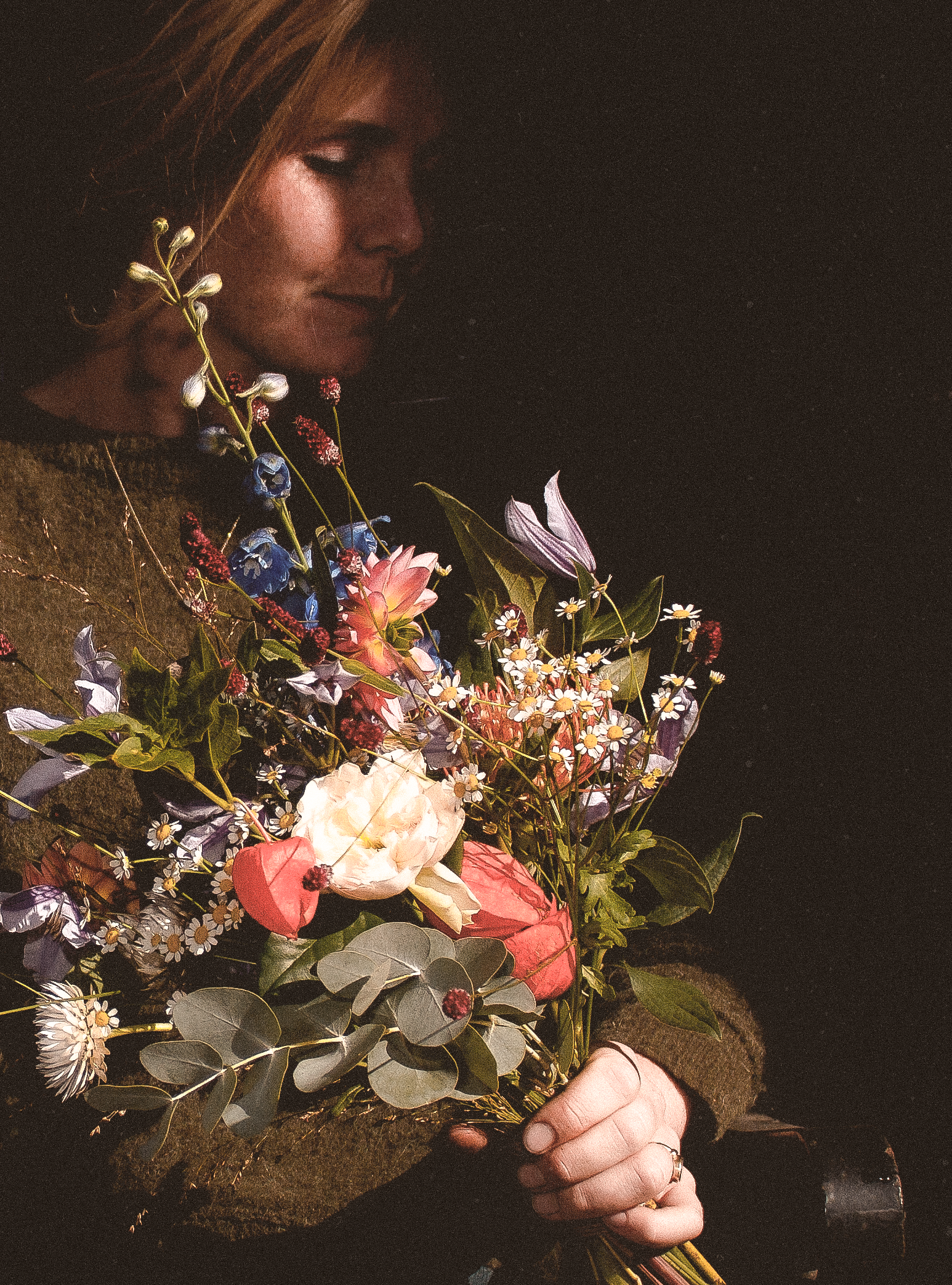 Persoon houdt een bos bloemen in een hand, de foto is genomen in een donkere setting met de persoon op de linkerzijde en een donkere achtergrond.