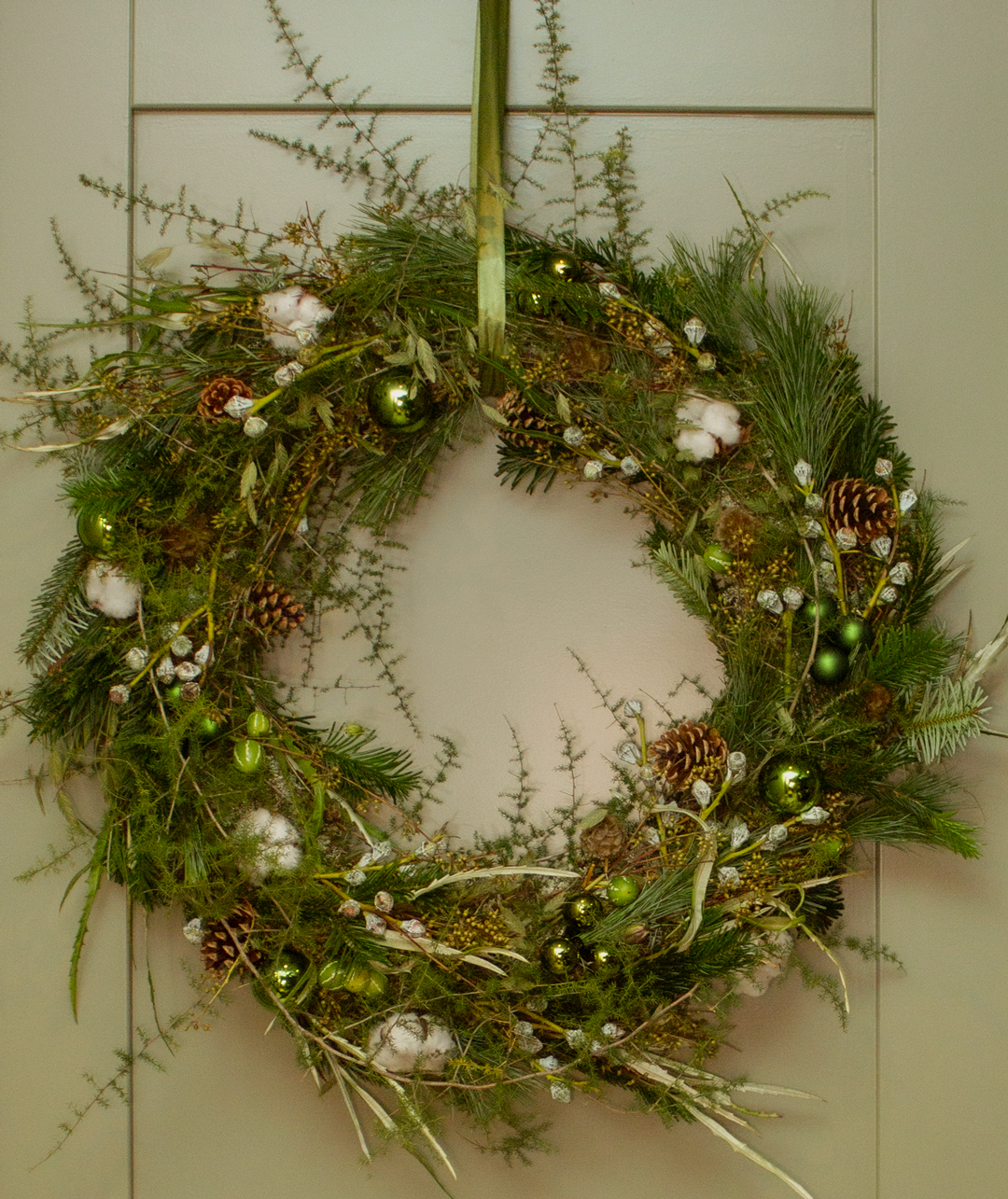 Een kerstkrans gemaakt van dennengroen, takken en decoraties zoals dennenappels, zilveren takjes en groene kerstballen, hangend aan een witte deur.