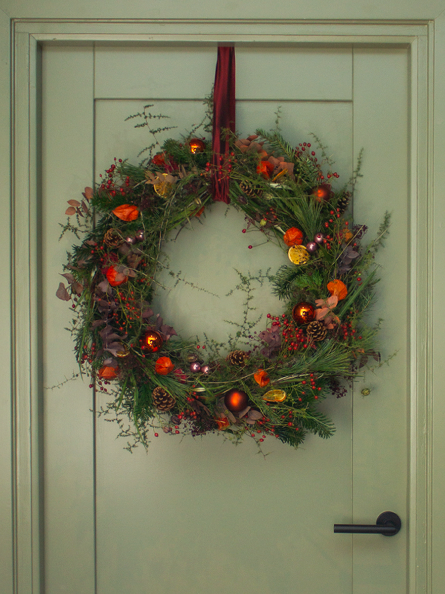 Kerstkrans hangend aan een deur met een rode lint in het midden, versierd met dennenappels, ballen, bloemen en groen.