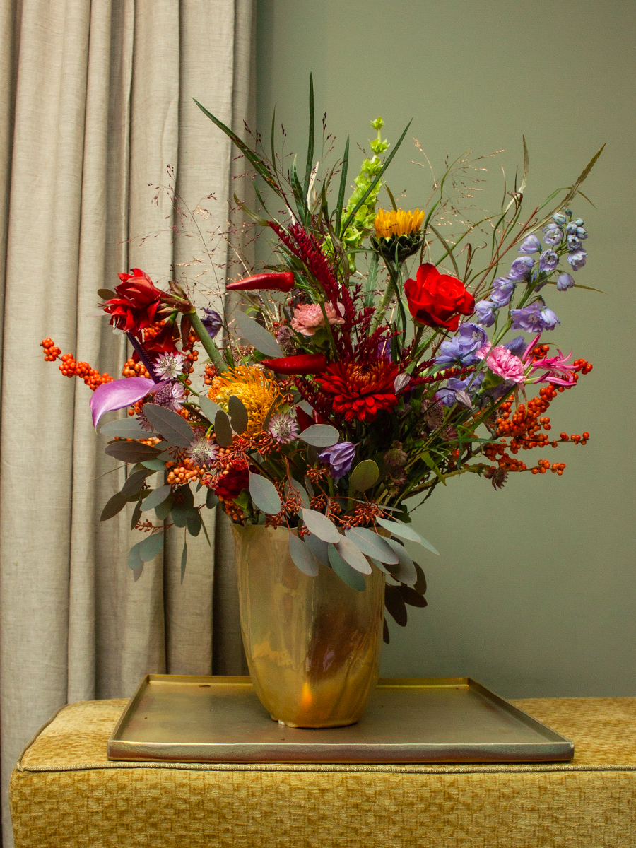 Kunstbloemen in een goudkleurige vaas op een tafel met een gouden dienblad, voor een beige gordijn en een groene muur.