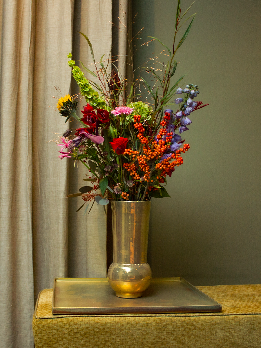 Een hoge, goudkleurige vaas met een kleurrijk boeket bloemen en groene bladeren, geplaatst op een tafel met een metalen dienblad, naast beige gordijnen en een groen muurtje.