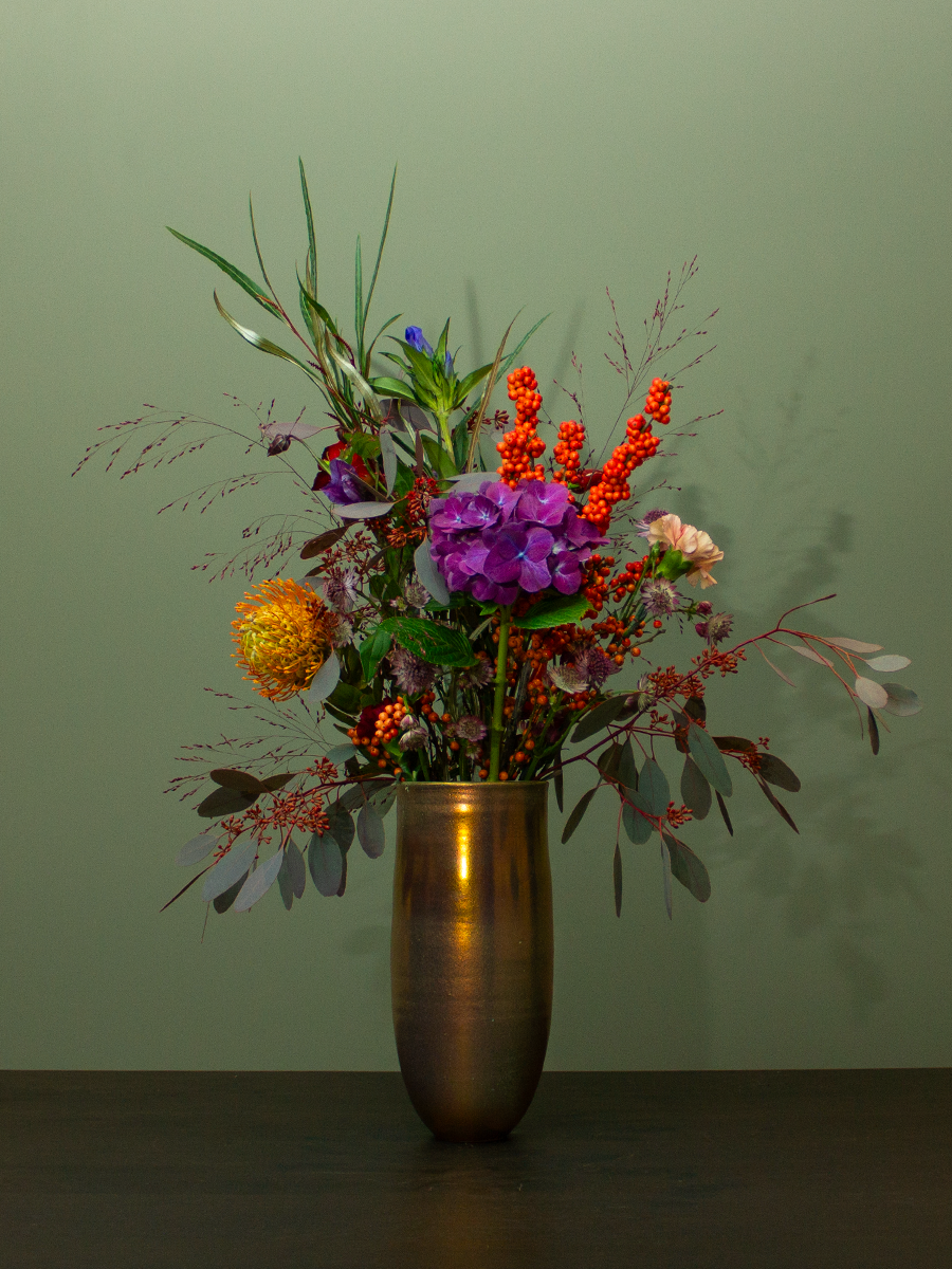 Een vaas met een kleurrijk boeket bloemen op een donkere tafel, tegen een lichtgroene achtergrond.