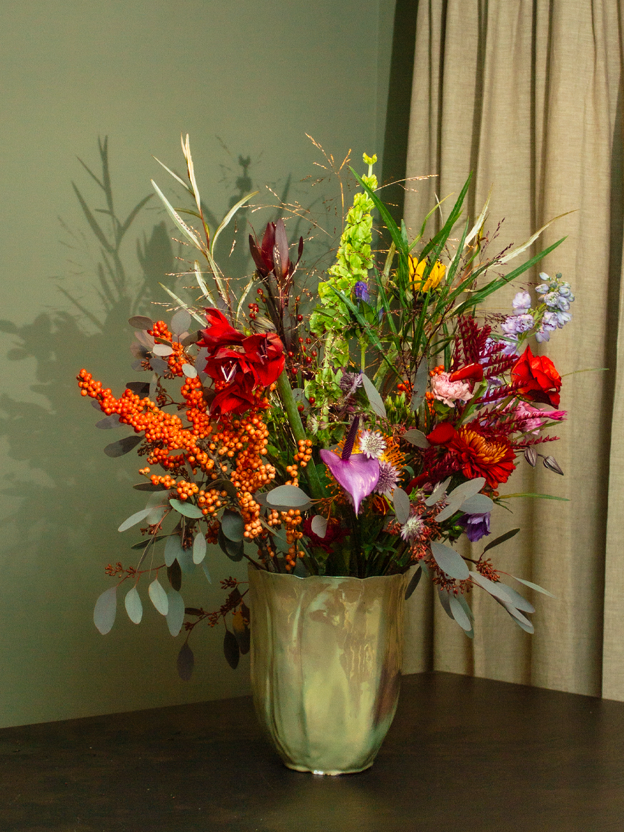 Een vaas met een kleurrijk boeket van bloemen en takken op een tafel, met een groene muur en beige gordijnen op de achtergrond.