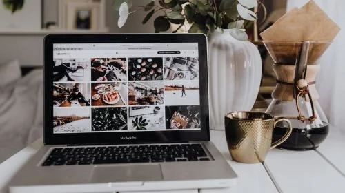 A laptop displaying a stock photo library, on a desk with a gold coffee cup, a jug of filtered coffee and a white vase filled with eucalyptus leaves.
