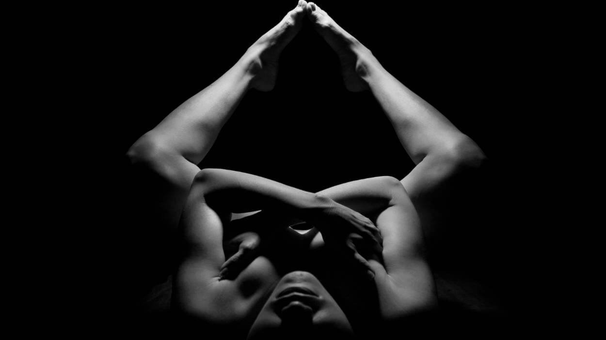 High-contrast, black and white fine art boudoir image of a person lying down with arms crossed over the chest and legs bent and raised, creating a powerful, symmetrical composition.