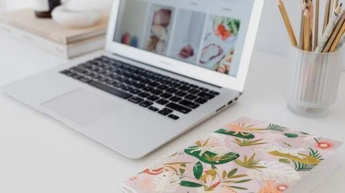 A chic laptop on a white desk, paired with styled stationery, a pot of pencils and a selection of books in the background.