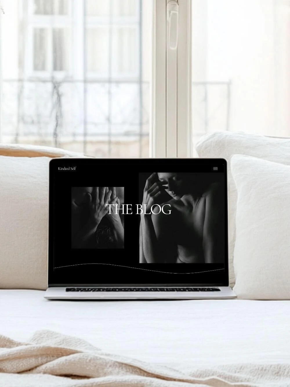Laptop displaying the “Kindred Spirit” website template’s blog page, showcasing two high-contrast boudoir images and the title text. The laptop is resting on a white sofa with a large window as a backdrop.