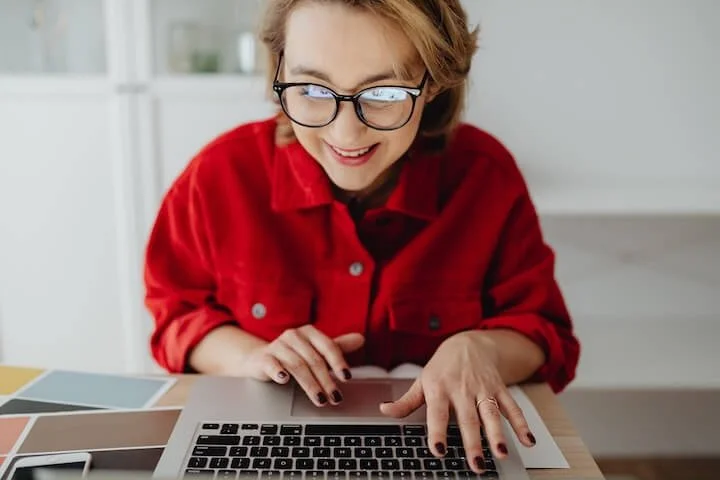 A smiling woman wearing glasses and a red jacket works on her laptop at a desk. Color swatches and a smartphone are placed on the table, suggesting a creative or design-related task