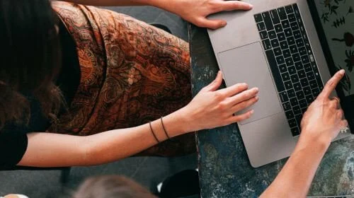 overhead shot of two women. One woman has her hands on the laptop and the other is pointing at the screen.