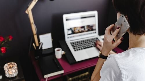 Woman, with back to the camera talking, on her mobile phone as she scrolls on her laptop whilst providing constructive feedback on the phone.