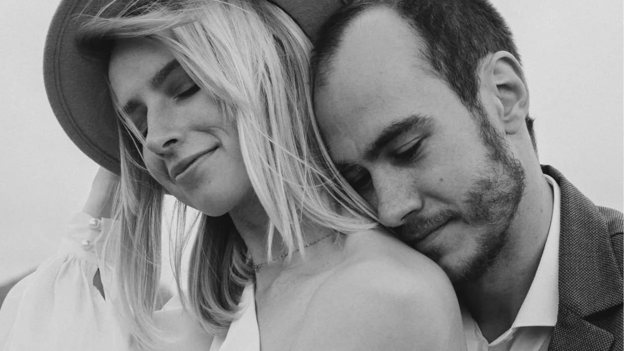 A black and white close-up portrait of a man resting his head on a woman's shoulder. The woman wears a hat and has her eyes closed, smiling gently. The man looks down, also with a gentle expression.