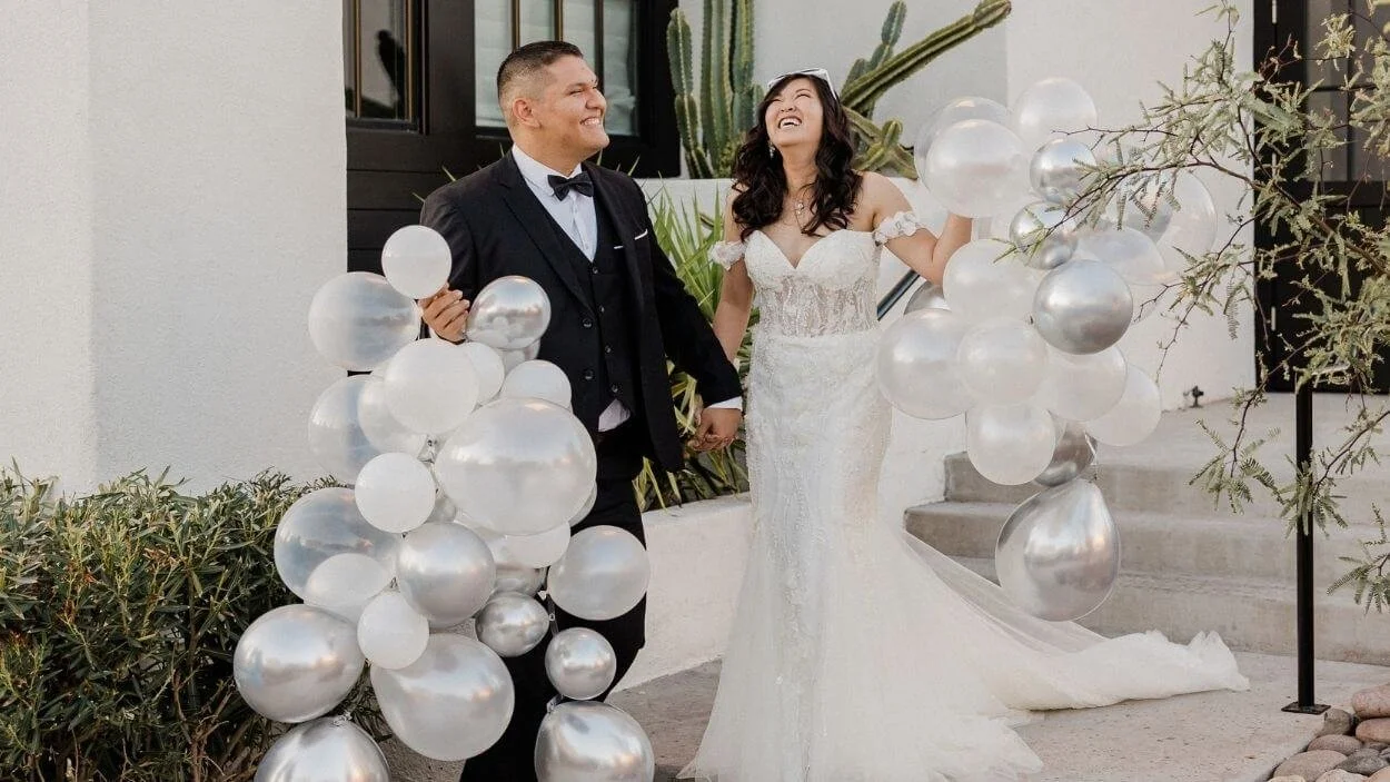 A full-length photo of a newly married couple holding hands and surrounded by white and silver balloons. The man is in a tuxedo and the woman is in a white wedding gown. They are both smiling and looking at the camera.