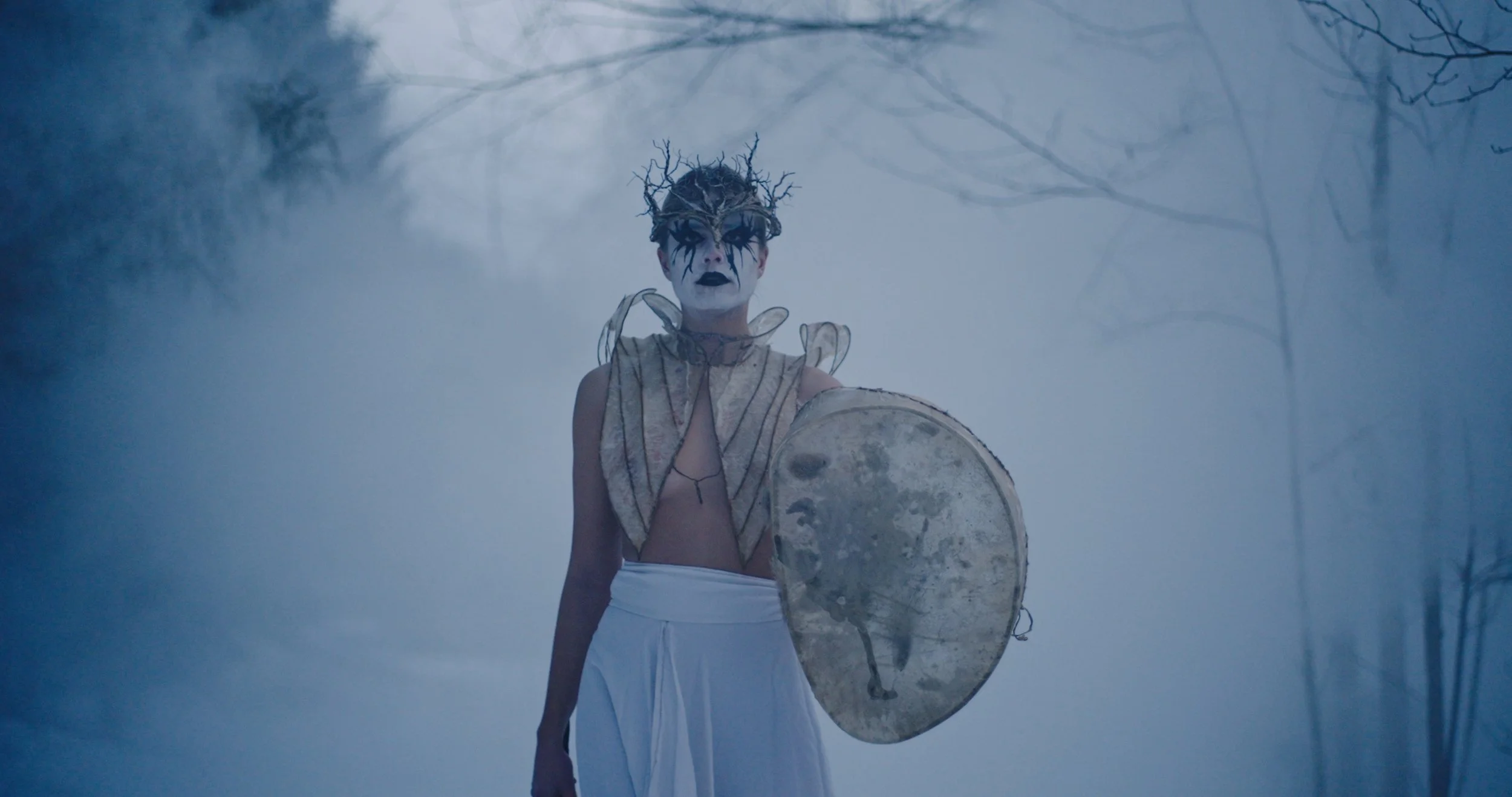 Stills bilde fra filmen "Hex". På bildet er det en kvinne som går i tåke. Kvinnen har malt ansiktet hvitt og svart rundt øyene. Leppene til kvinnen er svart. Det er Black-metal inspirasjon. Kvinnen har på en krone og går mot kamera med et skjold. 