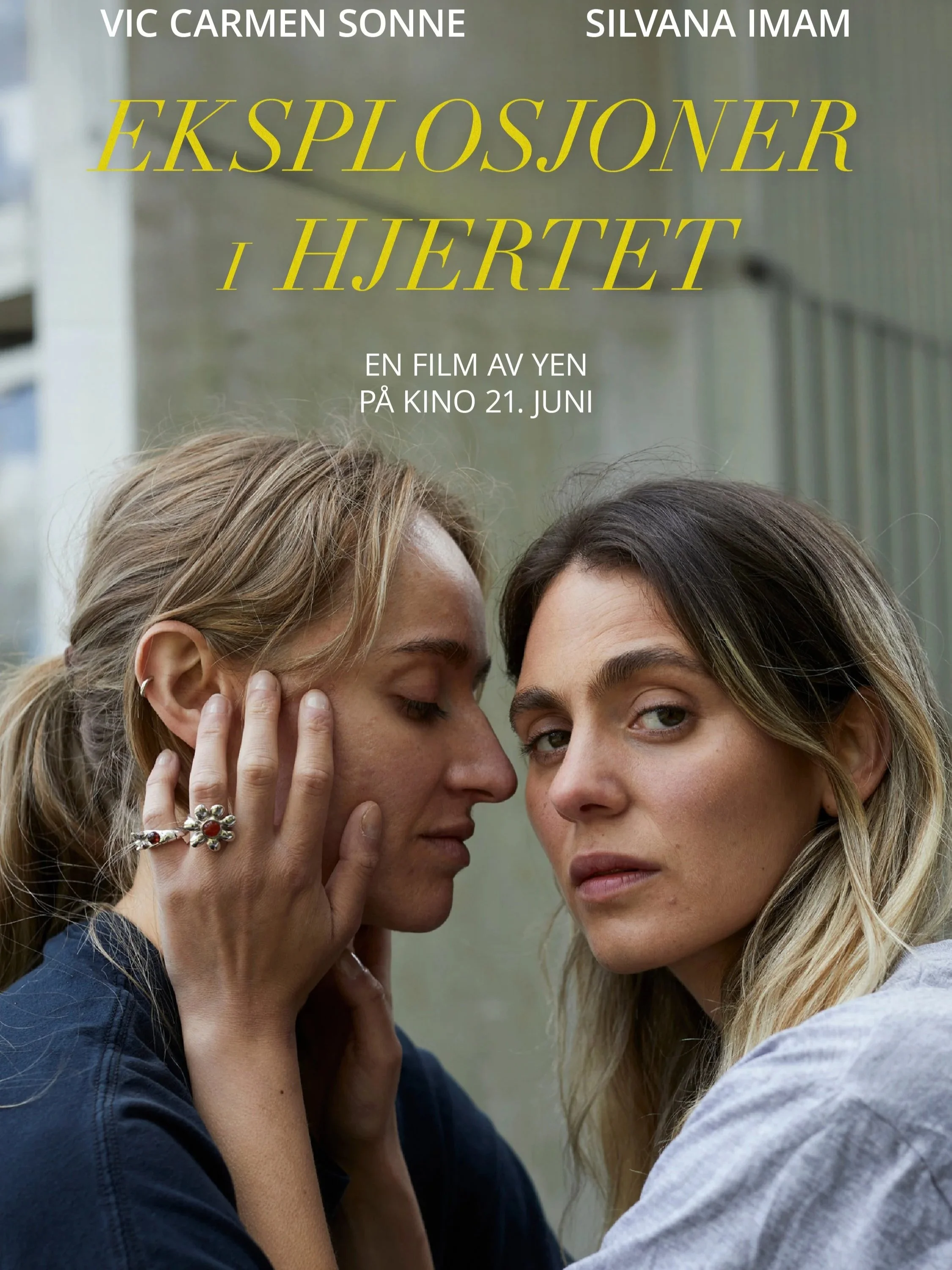 En filmplakat for filmen "Eksplosjoner i hjertet". Plakaten viser to jenter som står ansikt mot ansikt. Den ene jenta holder hendene kjærlig rundt ansiktet til hun andre mens hun ser inn i kamera. Den andre jenta lener inn i hendene hennes. Det er en
