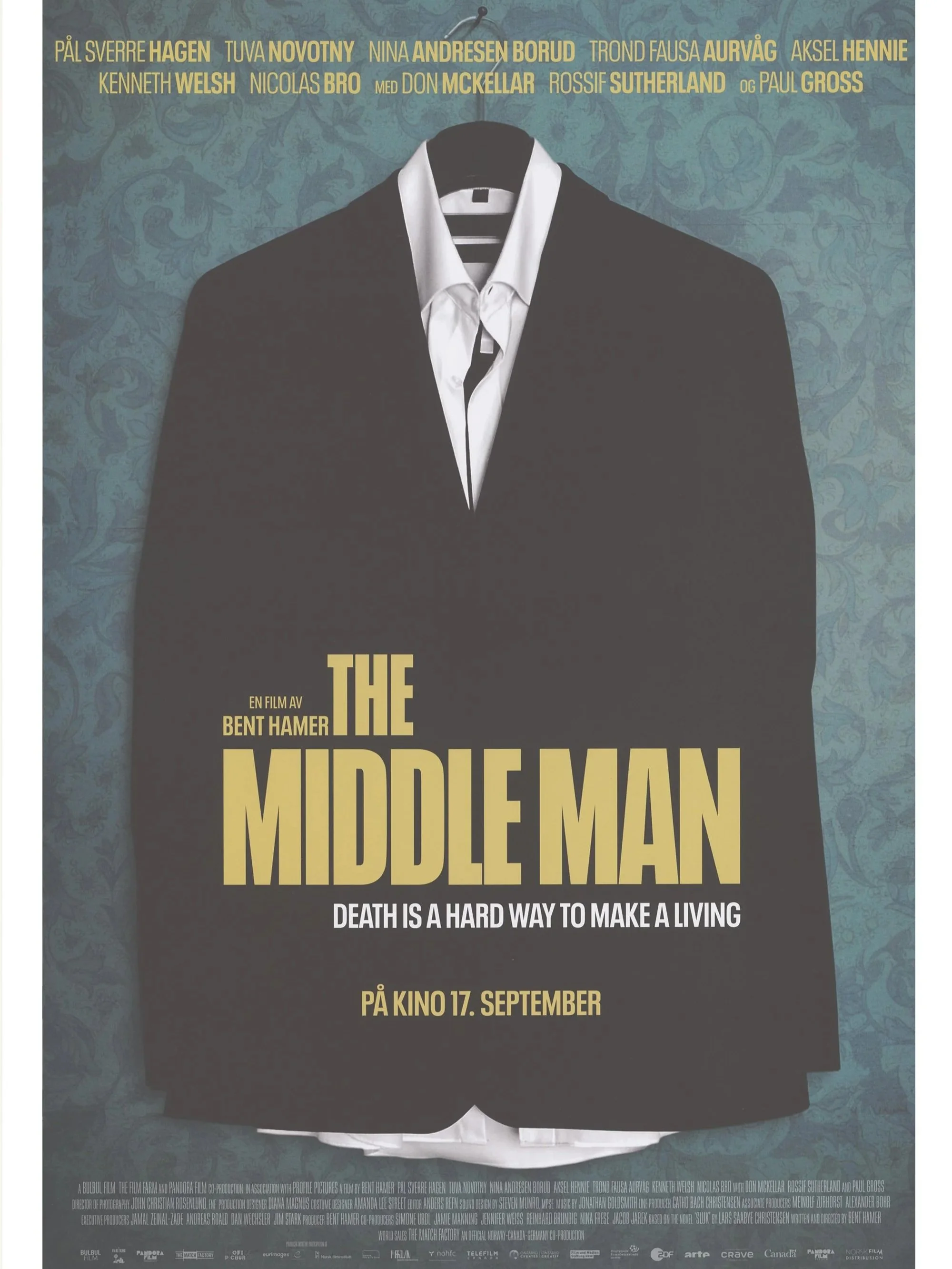 Filmplakat for "The middle man". Fokuset i plakaten er en svart dress med hvit skjorte som henger på en kleshenger. Dressen henger foran en grønn tapet med floralt mønster. Tittelen er midt på plakaten i gul skrift. 