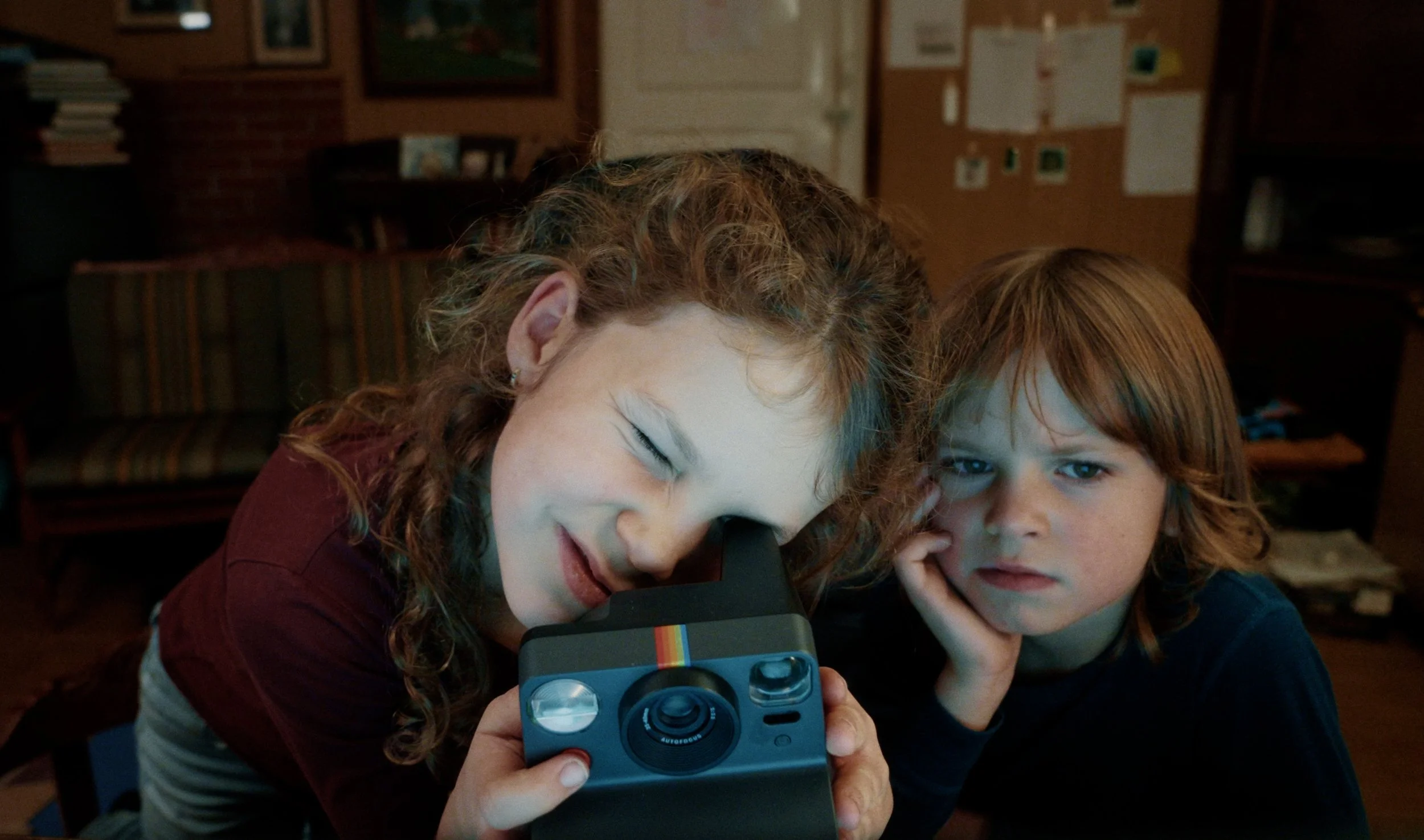 Stills bilde fra filmen "Mysteriepakken". På bildet er det to barn på alderen 5 og 8 år. Til høyre er det en liten gutt med litt langt hår som ser mot en skjerm. Til venstre er det en jente med krøllete hår som ser i et digitalt kamera.