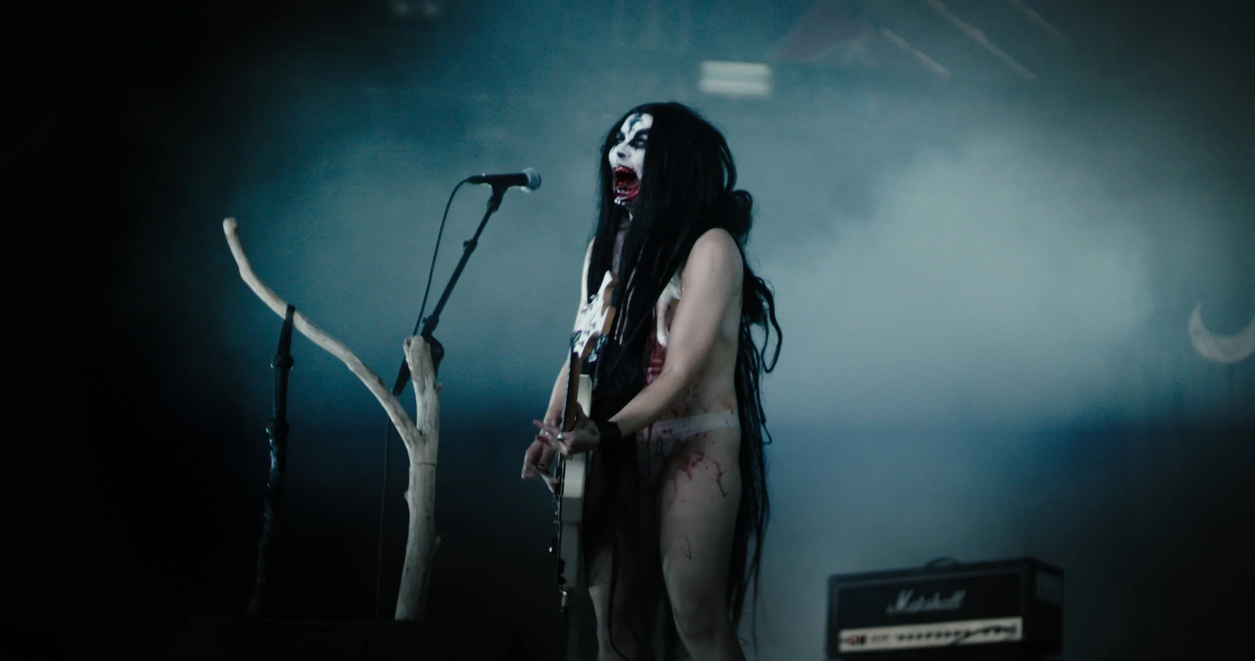 Stills bilde fra filmen "HEX". Det er en black-metal kvinne som står på en scene i undertøy. Hun står foran mikrofonstativet og holder en gitar. Kvinnen har langt svart hår, og har blod på kroppen sin. Kvinnen skriker ut mot publikum.