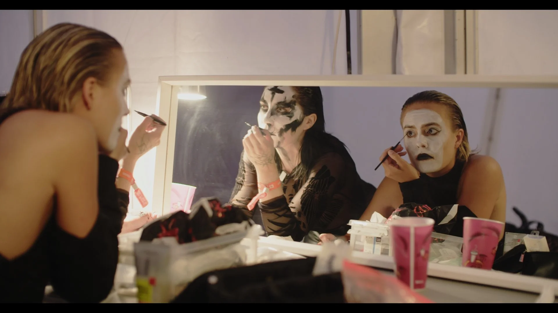 Stills bilde fra filmen "HEX". På bildet er det to kvinner som sminker seg foran et speil. Kvinnene sminker seg med hvit og svart maling i ansiktet. 