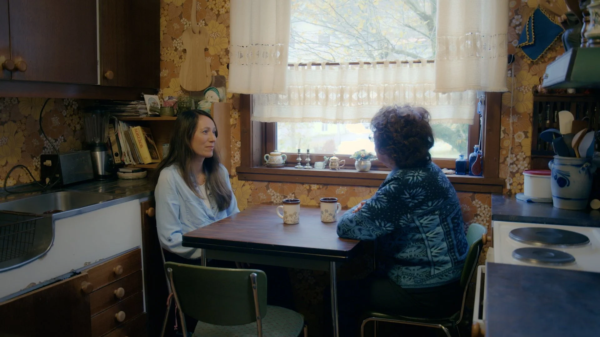 Stills bilde fra dokumentaren "De rettsløse". På bildet er det en yngre og en eldre kvinne som sitter rundt et lite spisebord. Kjøkkenet ser eldre ut, typisk 70-års tallet. 