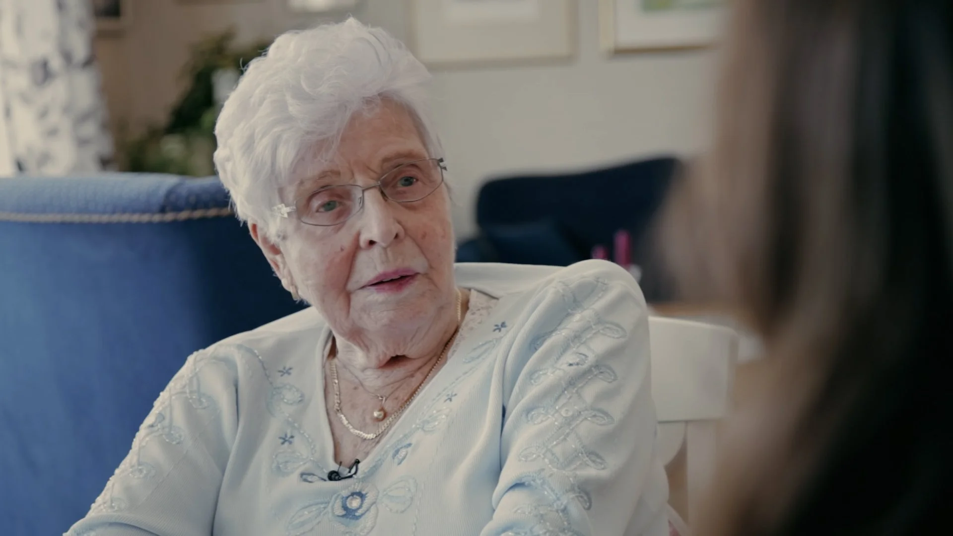Stills bilde fra dokumentaren "De rettsløse". På bildet er det en gammel kvinne som prater med noen. Hun har grått, kort hår og har på en lyseblå bluse. 
