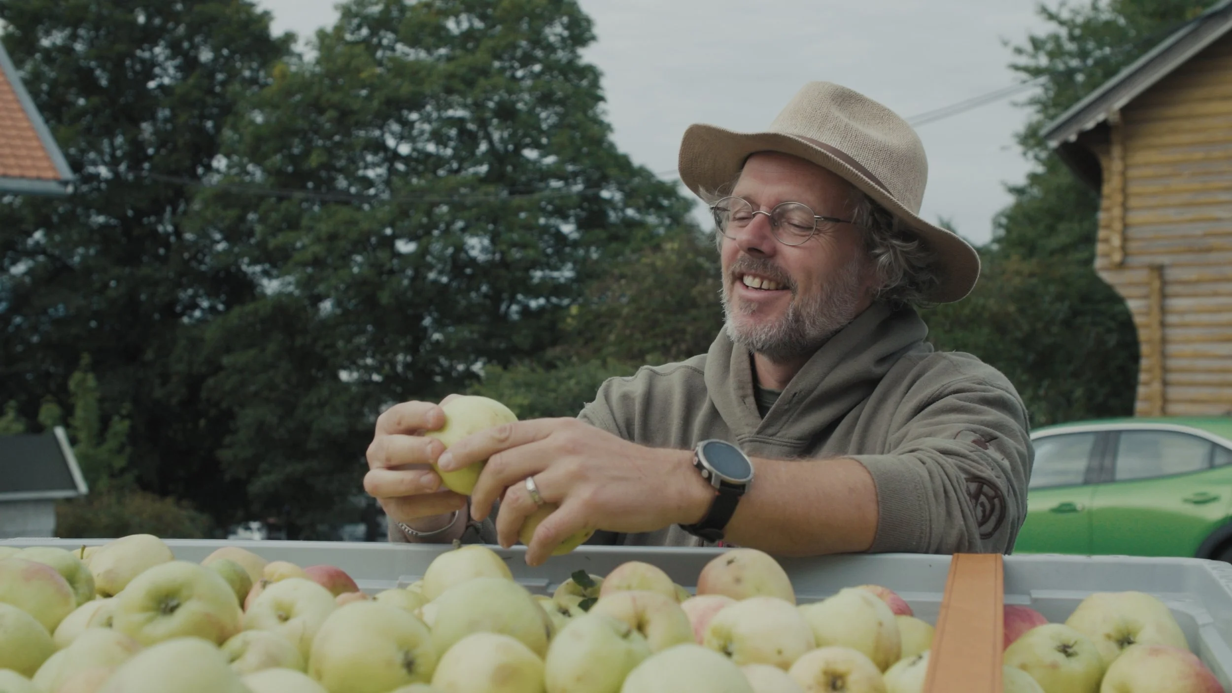Stills bilde fra filmen "Dette er ikke et eple". På bildet er det en mann med en hatt som smiler ned mot et eple. Foran mannen er det en kasse full av grønne og røde epler. 