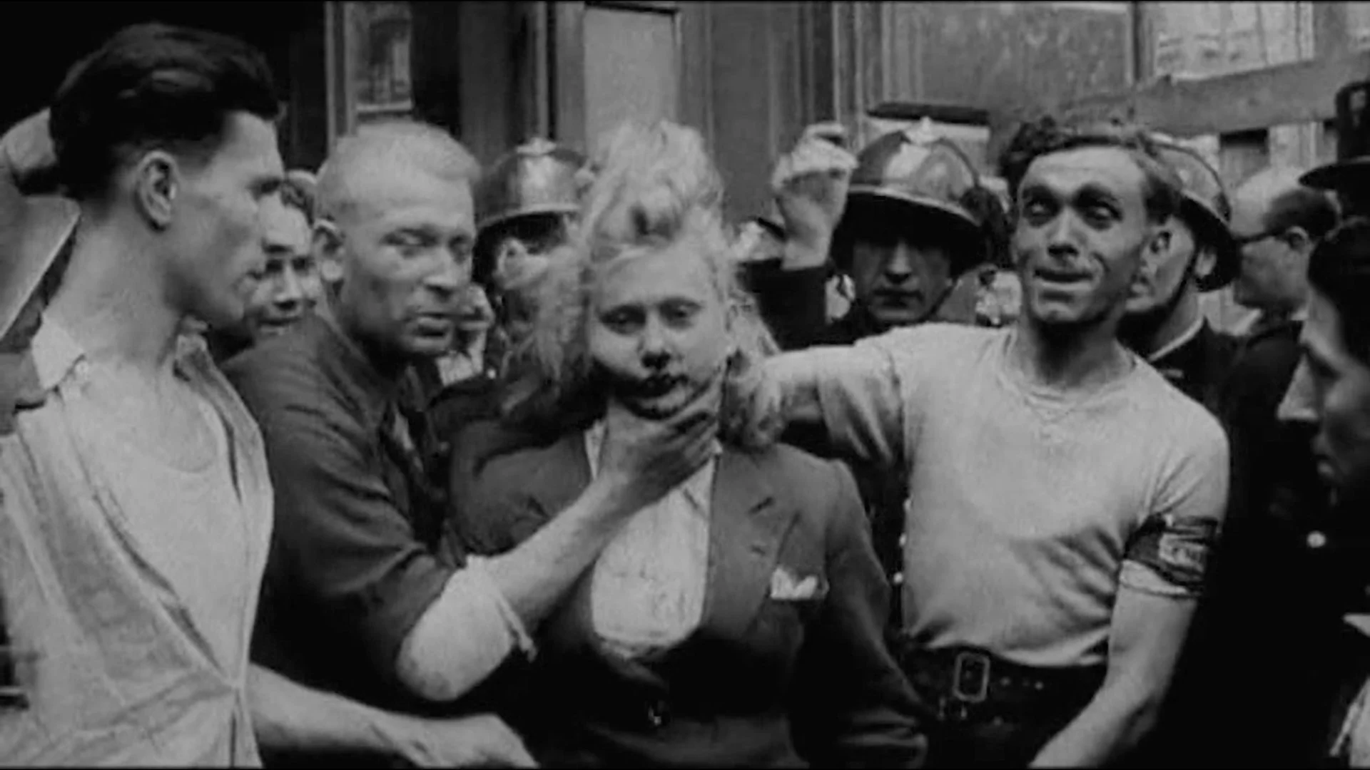 Still bilde fra filmen "De rettsløse". På bildet er det en kvinne fra andre verdenskrig som blir latterliggjort. Rundt kvinnen er det flere menn som ler og tar på kvinnen. Bildet er i svart og hvitt. 