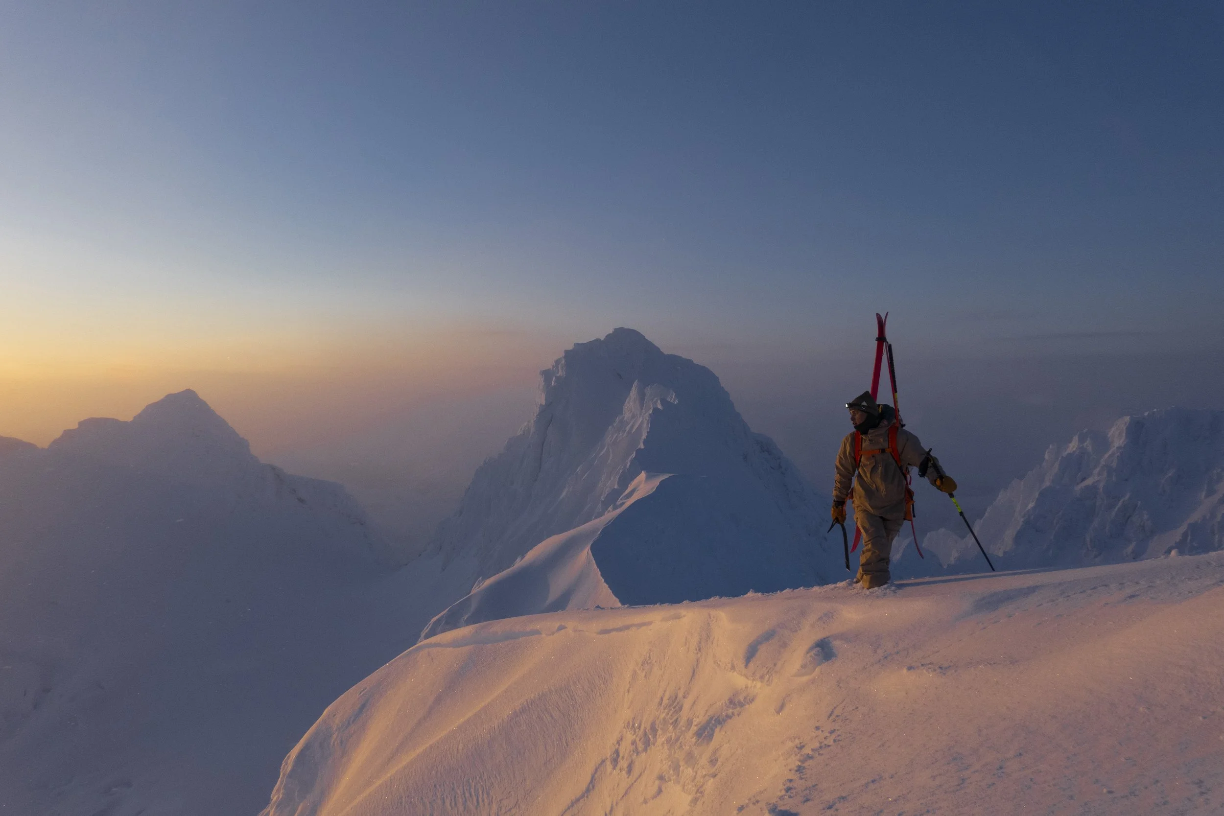 Stillsbilde fra filmen "SKI". En mann står på toppen av en fjelltopp og ser utover horisonten. I bakgrunnen er det flere fjelltopper og snø. Det er solnedgang, og snøen ser nesten gul ut. 