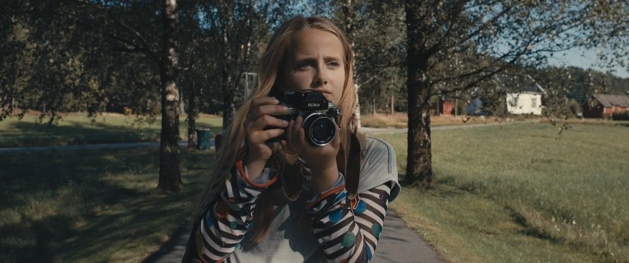 Et bilde av en blond jente på en gangsti. Hun står midt i bildet og holder et kamera som om hun er i ferd med å ta bilde av noe.
