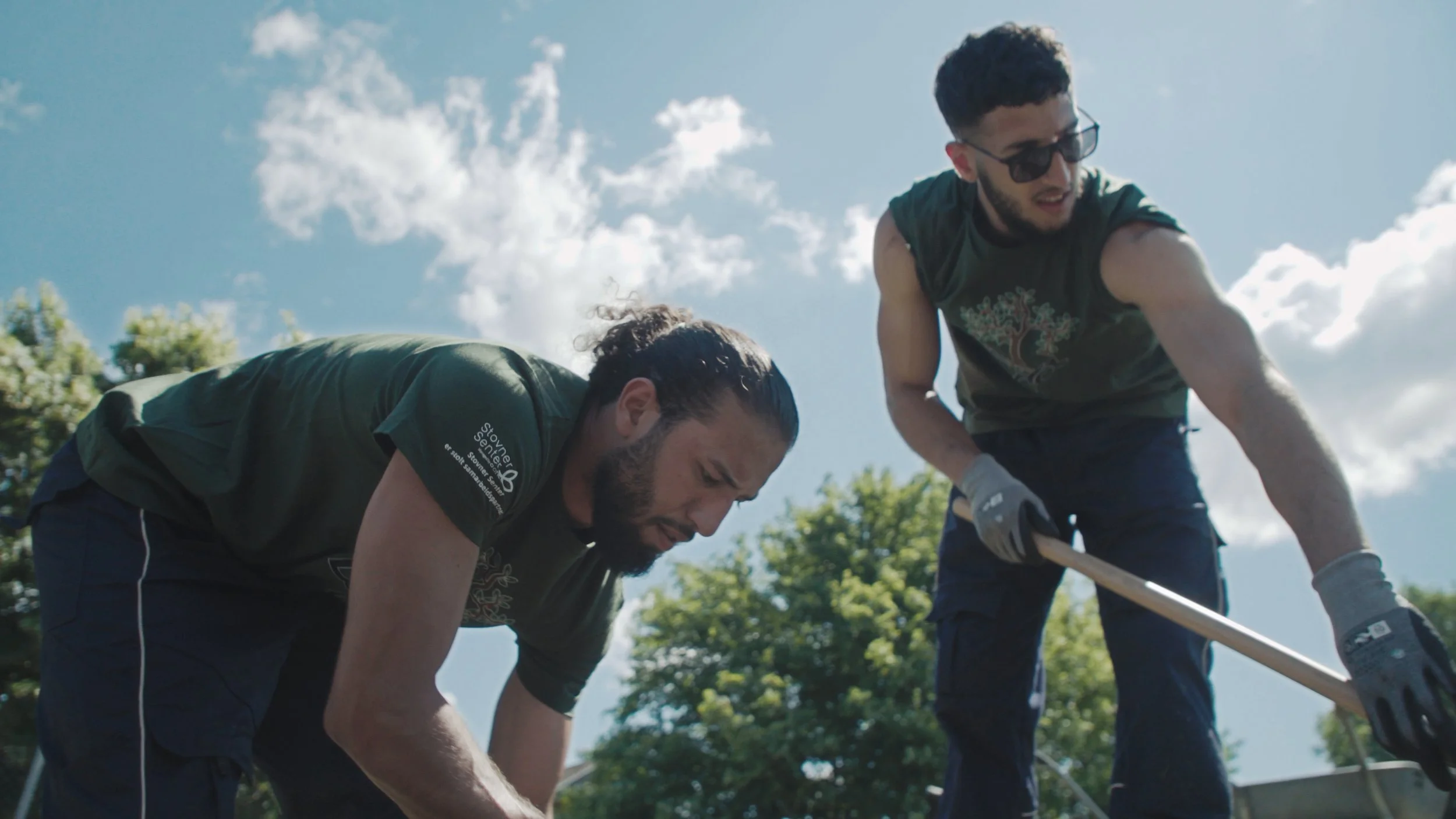 Still bilde av to karakterer fra filmen "Dette er ikke et eple", to menn som jobber for Lif Laga. Mannen til høyre har på solbriller og har mørkt, kort hår. Mannen til venstre har håret i en "man bin" og det ser ut som han graver etter noe. Bakgrunne